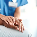 Photo of a medical professional in blue holding the hand of another person.