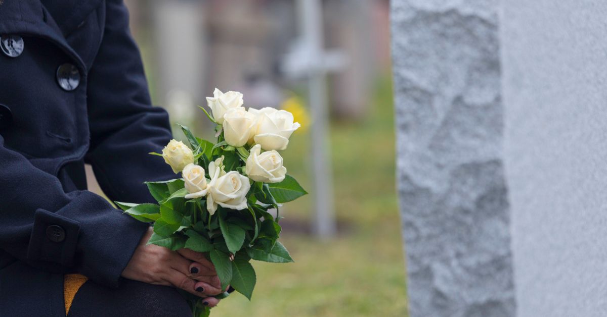 Woman kneeling with white roses before a headstone.